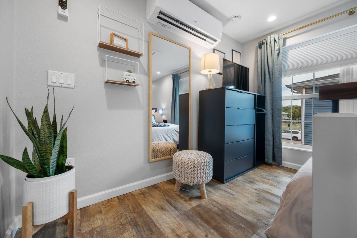 Une chambre d’hôtel moderne, équipée d’un lit, d’une armoire, d’un miroir, d’une plante en pot et d’une fenêtre avec des rideaux.