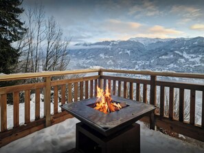 Una terraza con una mesa de fuego metálica rodeada de nieve, con vistas a un paisaje montañoso.