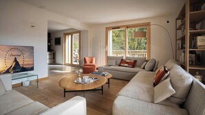 Un salon moderne avec un canapé, un fauteuil et une table basse. On y trouve également une bibliothèque et un téléviseur fixé au mur.