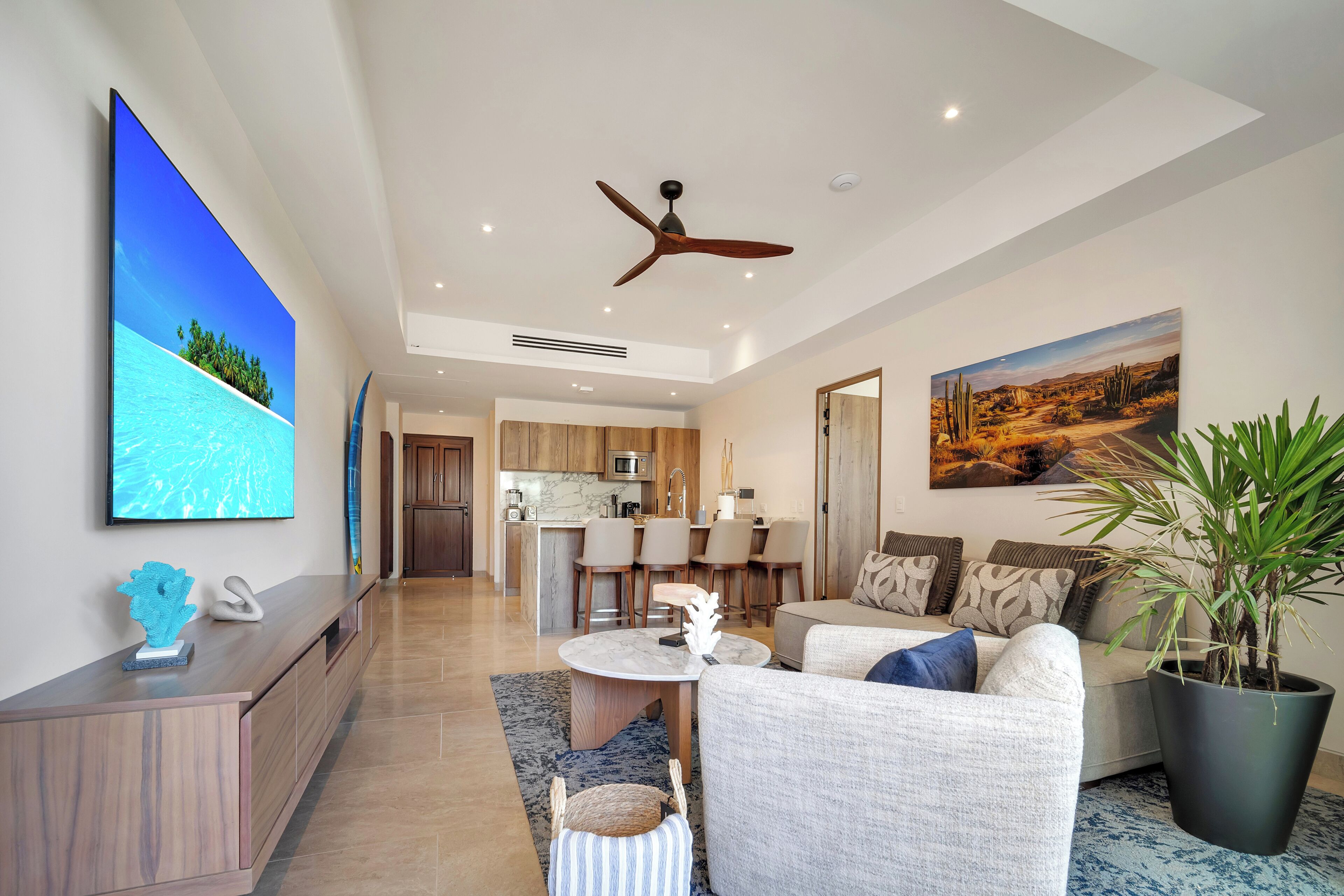 A modern living room with a flat-screen TV, a ceiling fan, a dining area, and a kitchen in the background.
