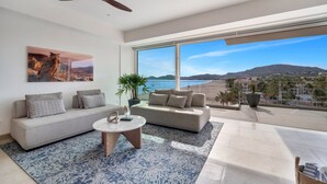 A modern living room with a large window, a ceiling fan, a sofa, a coffee table, and a potted plant. - Albaluz (San Jose del Cabo)