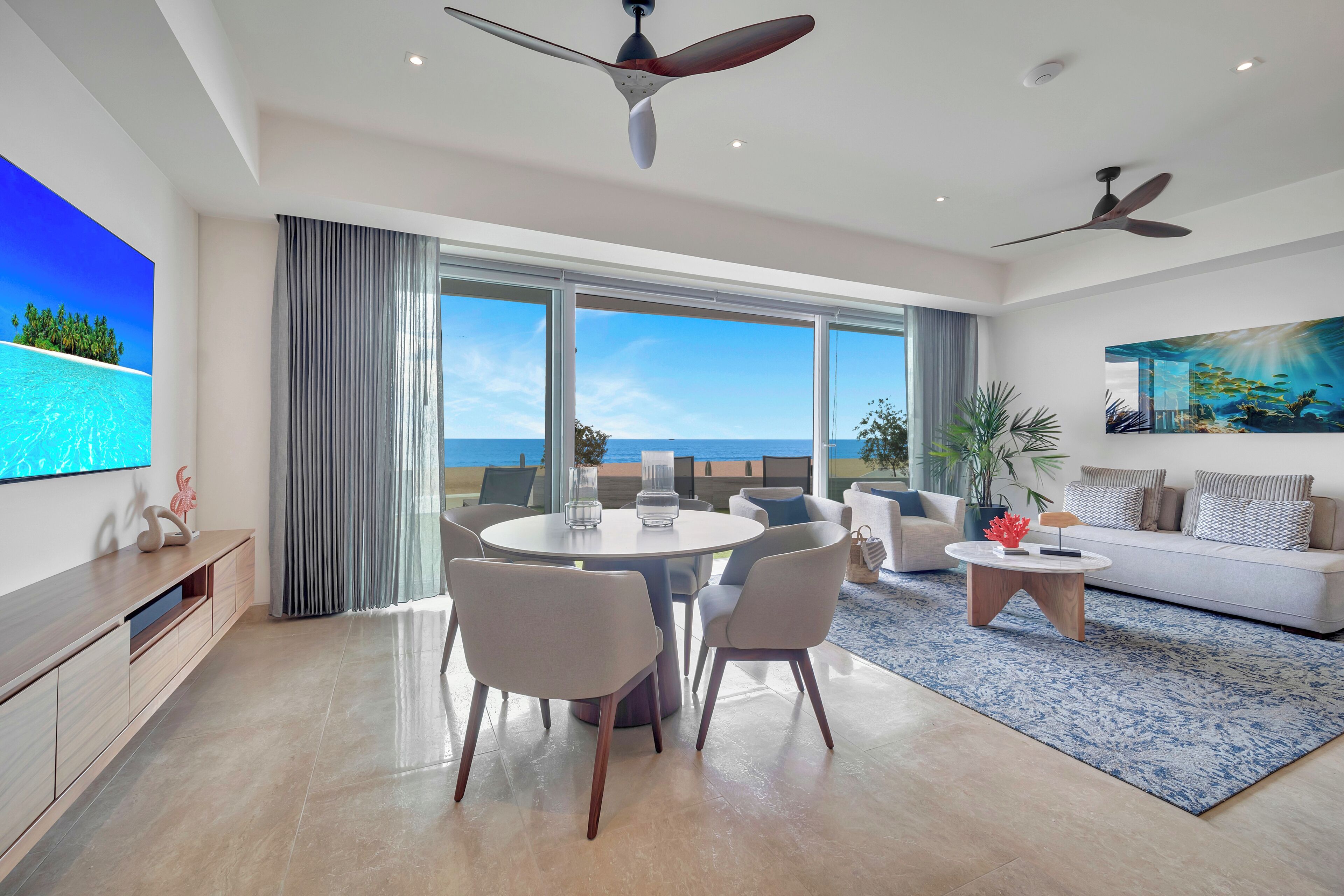 A modern living room with a dining area, large windows, ceiling fans, and a flat-screen TV.