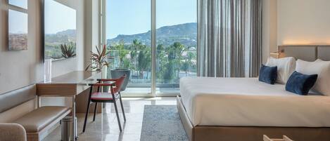 A modern hotel room with a large bed, a desk, a chair, and a view of the city and mountains.