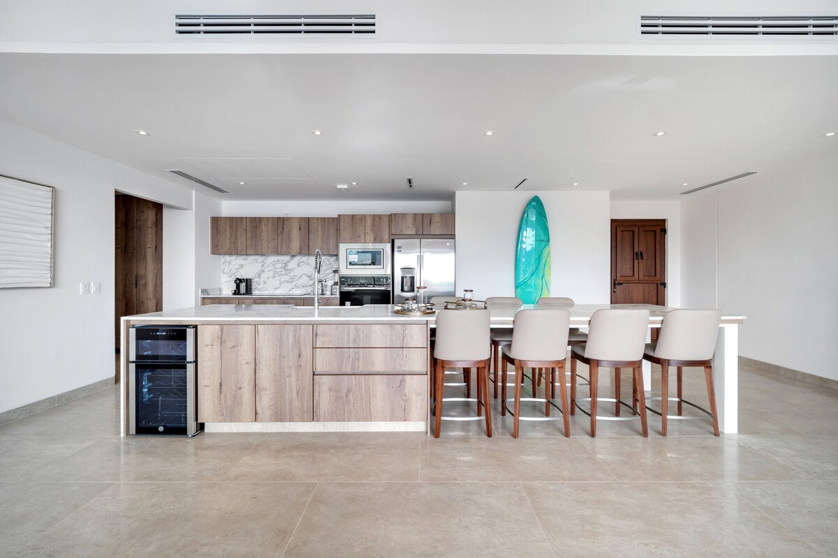 A modern kitchen with wooden cabinets, a marble backsplash, and a dining area with a large island and chairs.