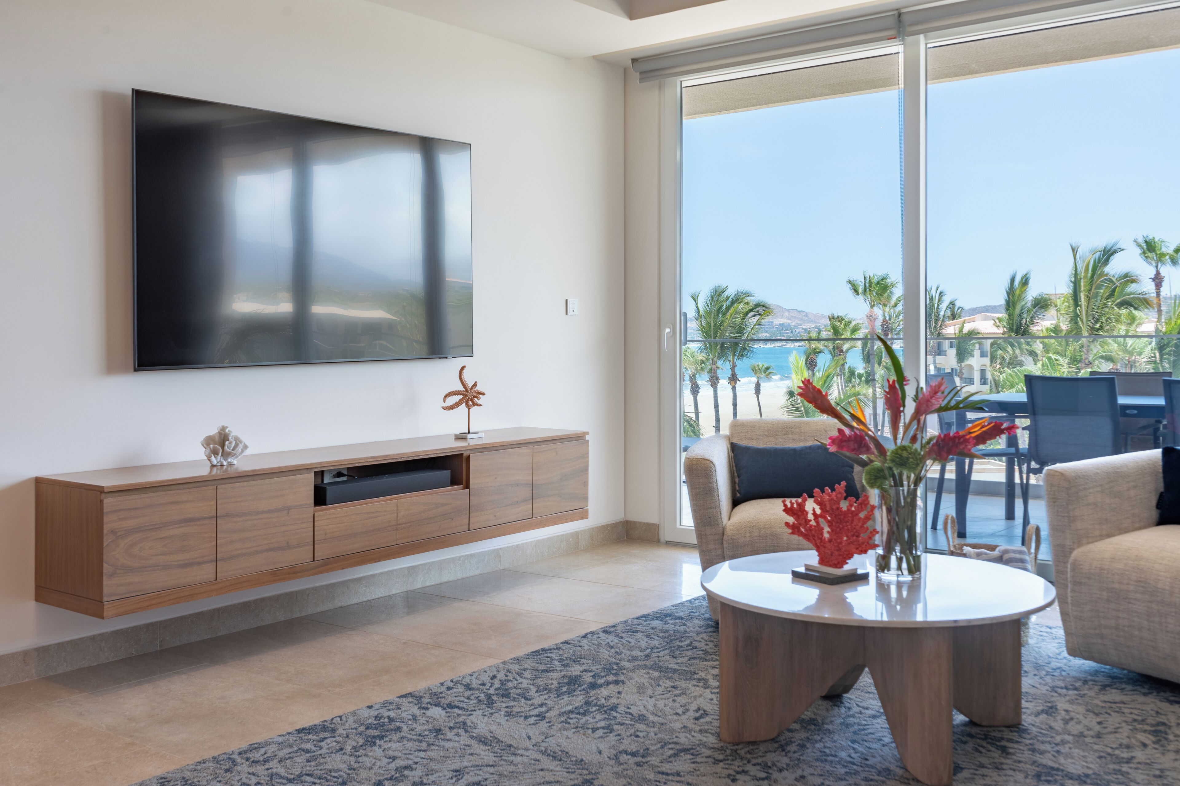 Una sala de estar moderna con un televisor grande, un mueble de madera, una mesa de centro redonda con un florero y vistas al océano a través de amplios ventanales correderas.