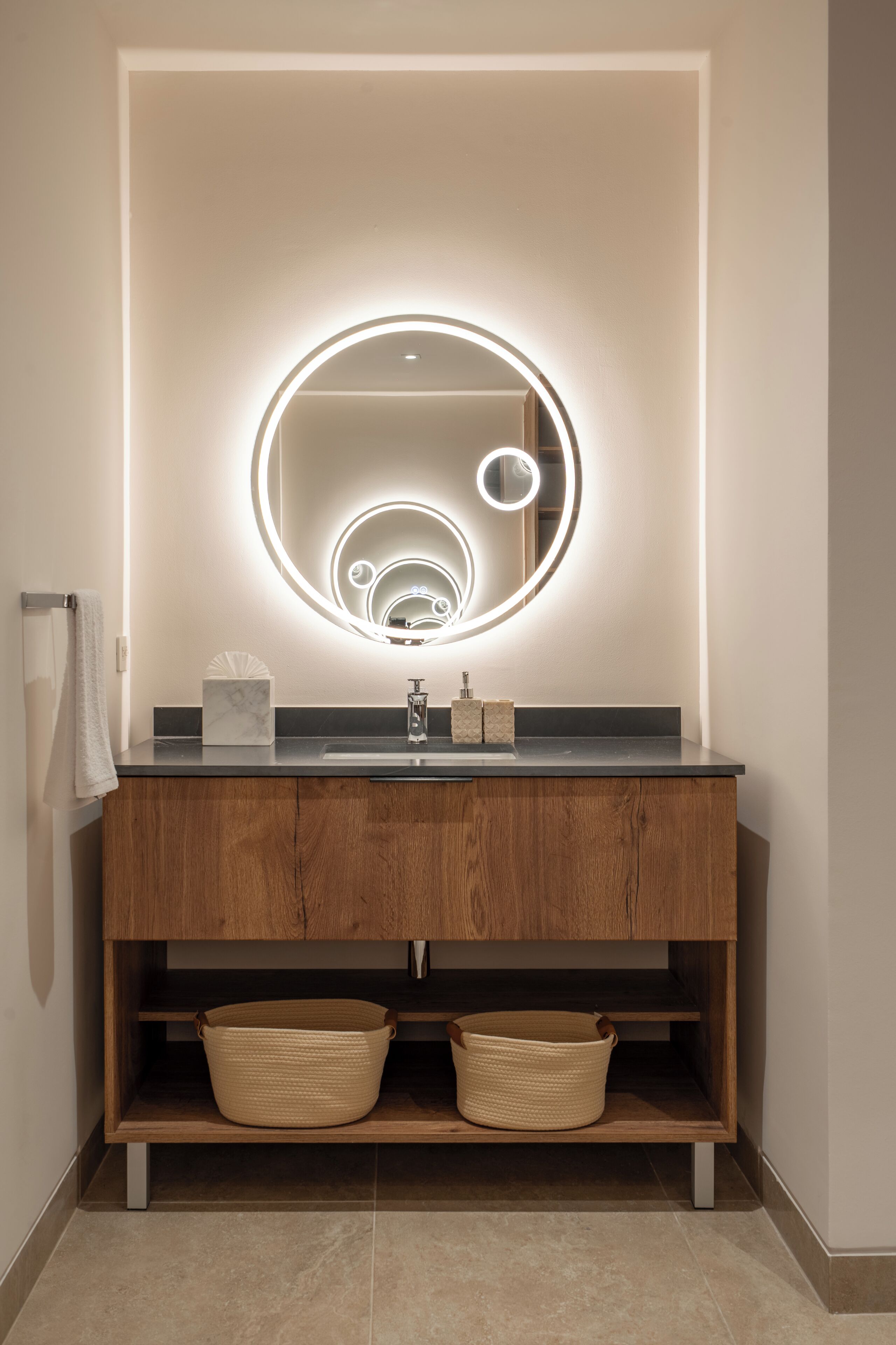 A bathroom with a wooden vanity, a round mirror with lights, a towel rack, and two baskets.