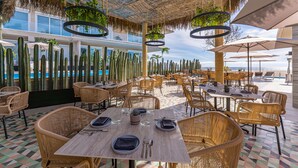 An outdoor dining area with wicker furniture, tables set with plates, cutlery, and glasses, and a view of the ocean. - Albaluz (San Jose del Cabo)