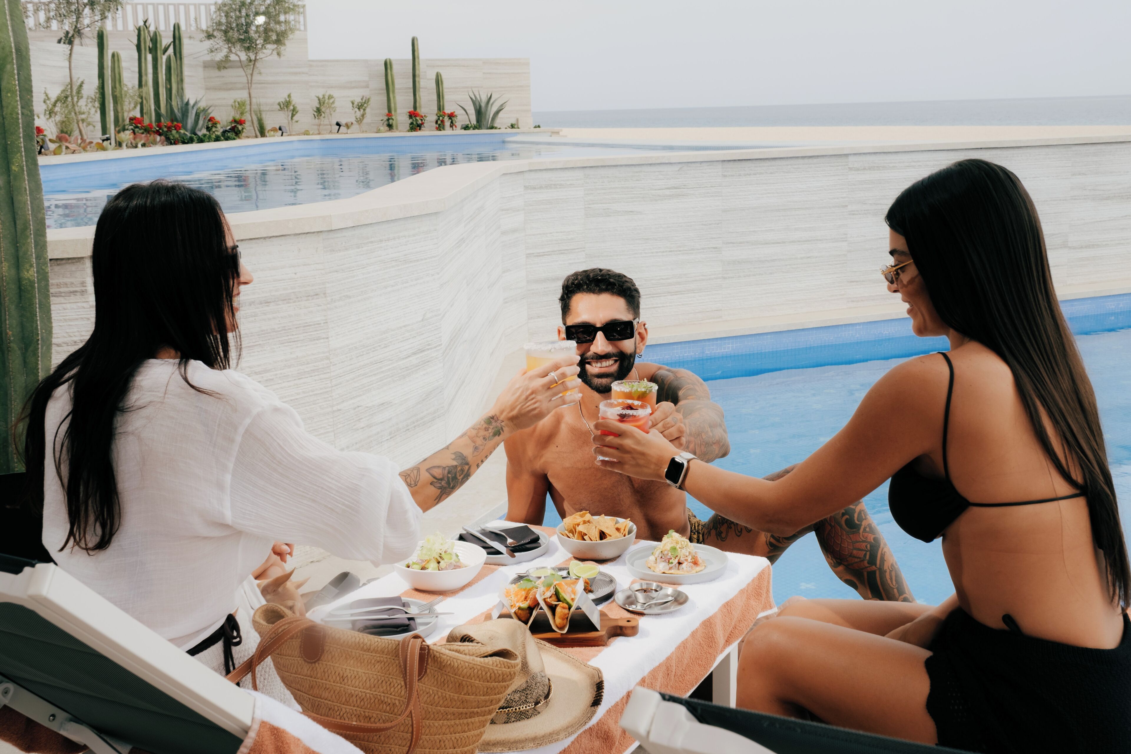 A poolside dining area with a table set for three, surrounded by cacti and a view of the ocean.