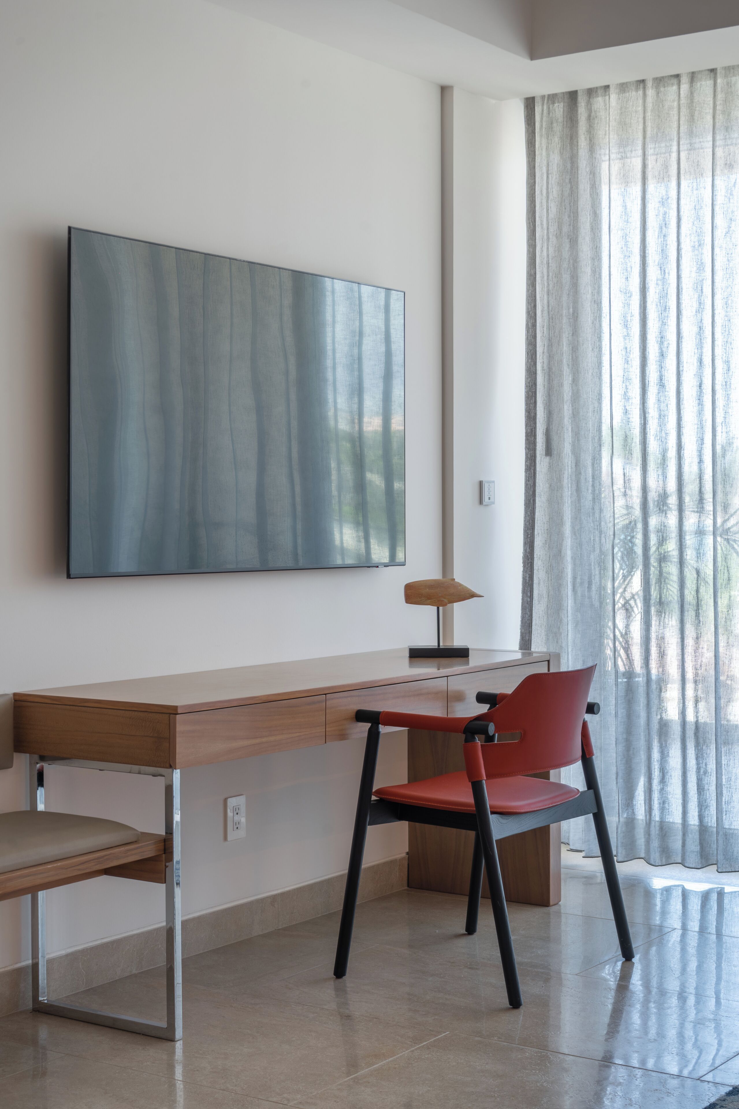 Una camera moderna con una scrivania in legno, una sedia rossa, un grande quadro astratto e una finestra con tende leggere.