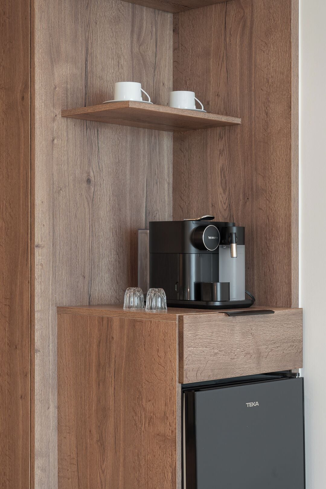 Una cocina con cafetera, dos tazas en un estante y un refrigerador.