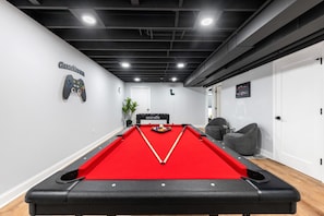 Una mesa de billar con superficie de fieltro rojo, una vara de billar y bolas de billar. En la pared hay un letrero que dice 'GAMEROOM'.