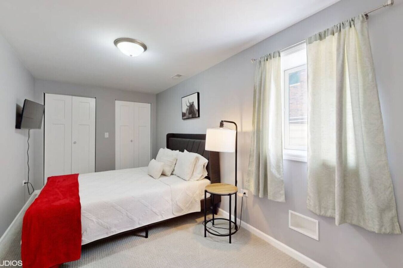 A bedroom with a bed, a bedside table, a lamp, a TV, and a window with curtains.