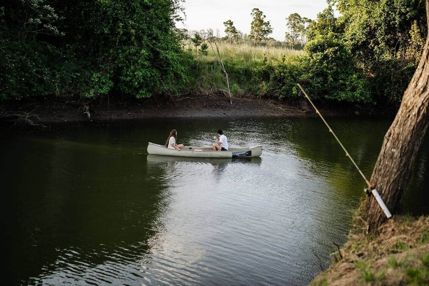 Une rivière paisible où une barque à rame transporte deux personnes, le tout entouré d’une végétation luxuriante.