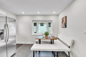 A compact kitchen with a built-in refrigerator, a dining table with a bench, and a window with blinds. - Bright & Cozy Elmhurst Stay • 3BR/2BA (Elmhurst)