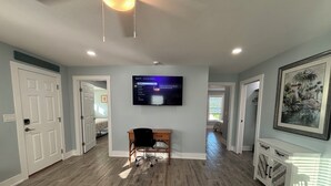 Een ruime woonkamer met houten vloeren, een wandgemonteerde flatscreen-tv, een houten bureau en een gekaderd schilderij aan de muur.