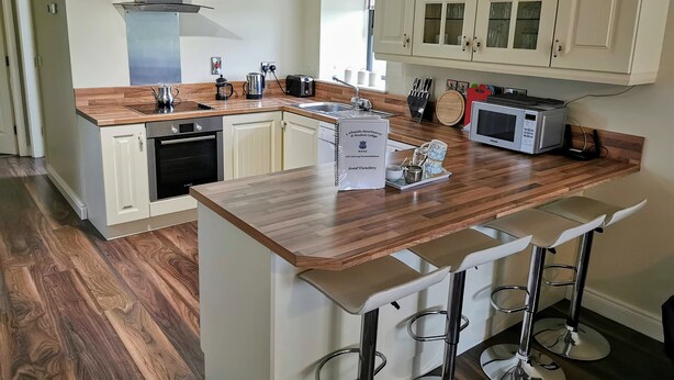 A modern kitchen with wooden countertops, a built-in oven, a microwave, and a coffee maker. There are bar stools and a sign indicating a guest directory.