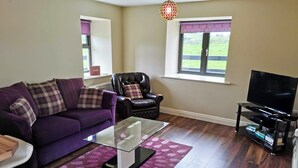 A living room with a purple sofa, a glass coffee table, a leather armchair, a TV on a stand, and a window with a view of greenery.