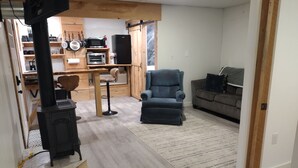 Una sala de estar acogedora con una chimenea moderna, un sillón cómodo, un sofá y una cocina con armarios de madera y varios electrodomésticos.
