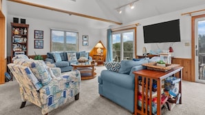 A spacious living room with a large sofa, armchairs, and a coffee table. There is a bookshelf, a TV mounted on the wall, and a balcony visible through the glass doors. - 3946 - Summers by the Sea - Duck OBX Retreat w/ Pool & Hot Tub Near Beach (Duck)