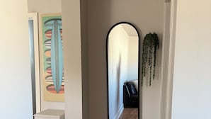 Un couloir avec un plancher en bois, un mur blanc, un miroir à cadre noir et une plante suspendue.