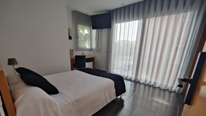 A hotel room with a bed, a desk, a chair, a TV, and a view of the outside through large windows.