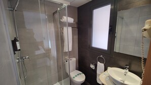 A modern bathroom with a glass-enclosed shower, a toilet with green symbols, a sink, and a window.