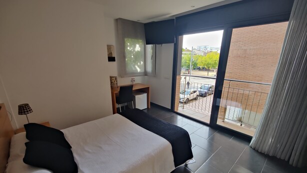 A modern hotel room with a large bed, a desk, and a view of the street through a sliding glass door.