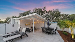 A covered outdoor patio with lounge chairs and a dining table, surrounded by a white fence and trees. - Sea La Vie | Salt Pool & built in Spa in great location (Sarasota)