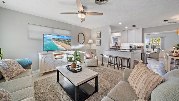A modern living room with a flat-screen TV, a ceiling fan, a dining area, and a kitchen in the background. - Sea La Vie | Salt Pool & built in Spa in great location (Sarasota)