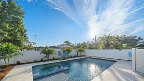 A rectangular swimming pool with a tiled deck, surrounded by a white fence, trees, and a clear blue sky. - Sea La Vie | Salt Pool & built in Spa in great location (Sarasota)