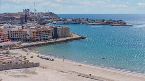 Eine Küstenstadt mit einem Hotel am Strand, einem felsigen Hügel im Hintergrund und einem klaren, blauen Meer.