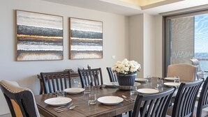 Un comedor con una mesa de madera dispuesta para seis personas, flanqueado por dos grandes cuadros abstractos, un jarrón de flores blancas y vistas al océano a través de un ventanal amplio.
