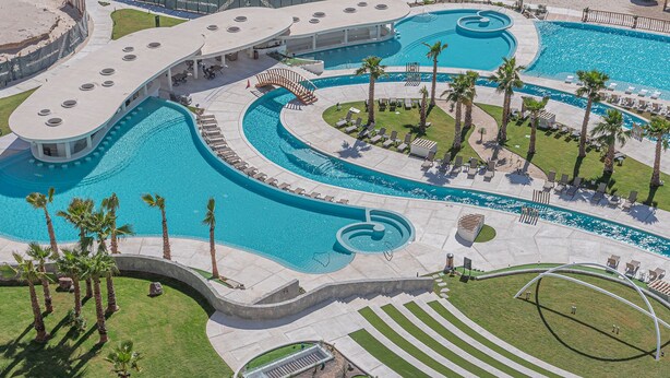 Una piscina grande, de varios niveles y formas diversas, con una terraza circundante, sillas de descanso y palmeras.