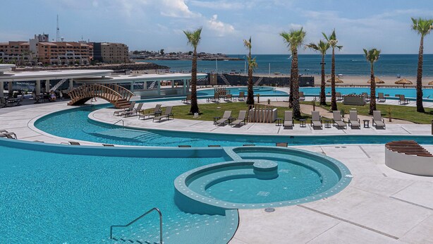 Ein Resort-Poolbereich mit mehreren Schwimmbecken, Liegestühlen, Palmen und Blick auf Strand und Meer.