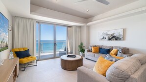 Amplia sala de estar con un gran ventanal corredizo que da a un balcón con vistas al mar. Hay un sillón amarillo, un sofá gris con cojines de colores y una mesa de centro de madera.