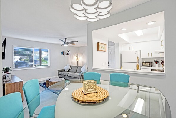 A modern living room with a glass dining table, teal chairs, a ceiling fan, and a view into the kitchen. - Pet Friendly Lauderdale by The Sea Beach Pad! (Lauderdale-by-the-Sea)