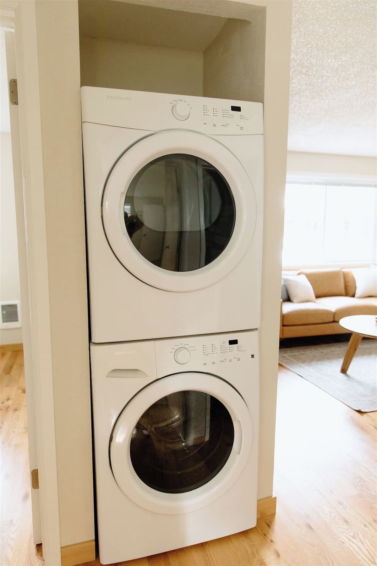 Une machine à laver et un sèche-linge superposés dans une pièce dont le sol est en bois et où l’on aperçoit un canapé beige à l’arrière-plan.