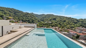 A rectangular pool with clear blue water, surrounded by a modern building with a flat roof and a view of a hillside with houses. - Casa Formosa · Modern 2BR + Rooftop Pool (Puerto Vallarta)