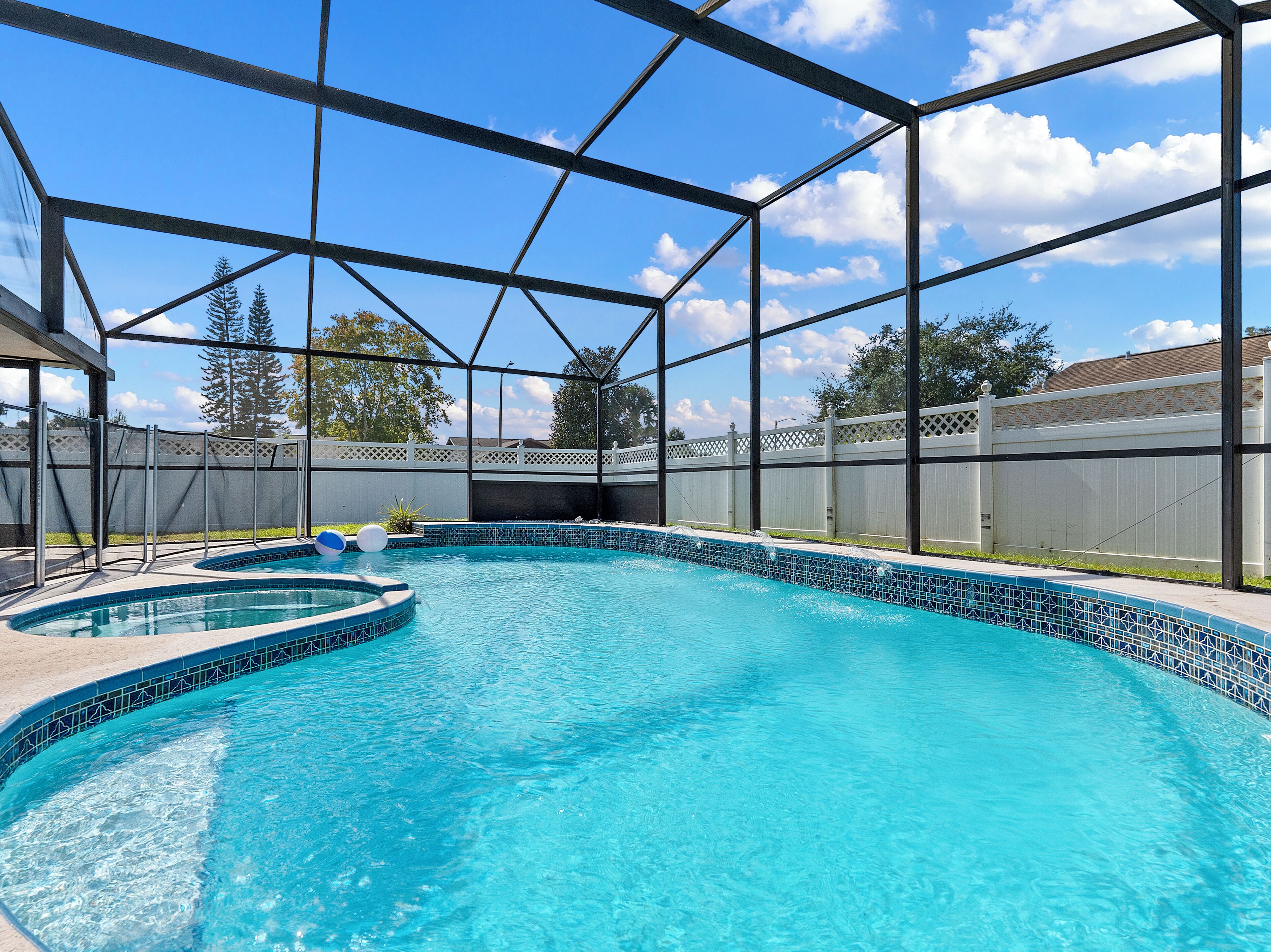 A swimming pool with a clear blue water, surrounded by a metal fence, and a view of trees and a house in the background.
