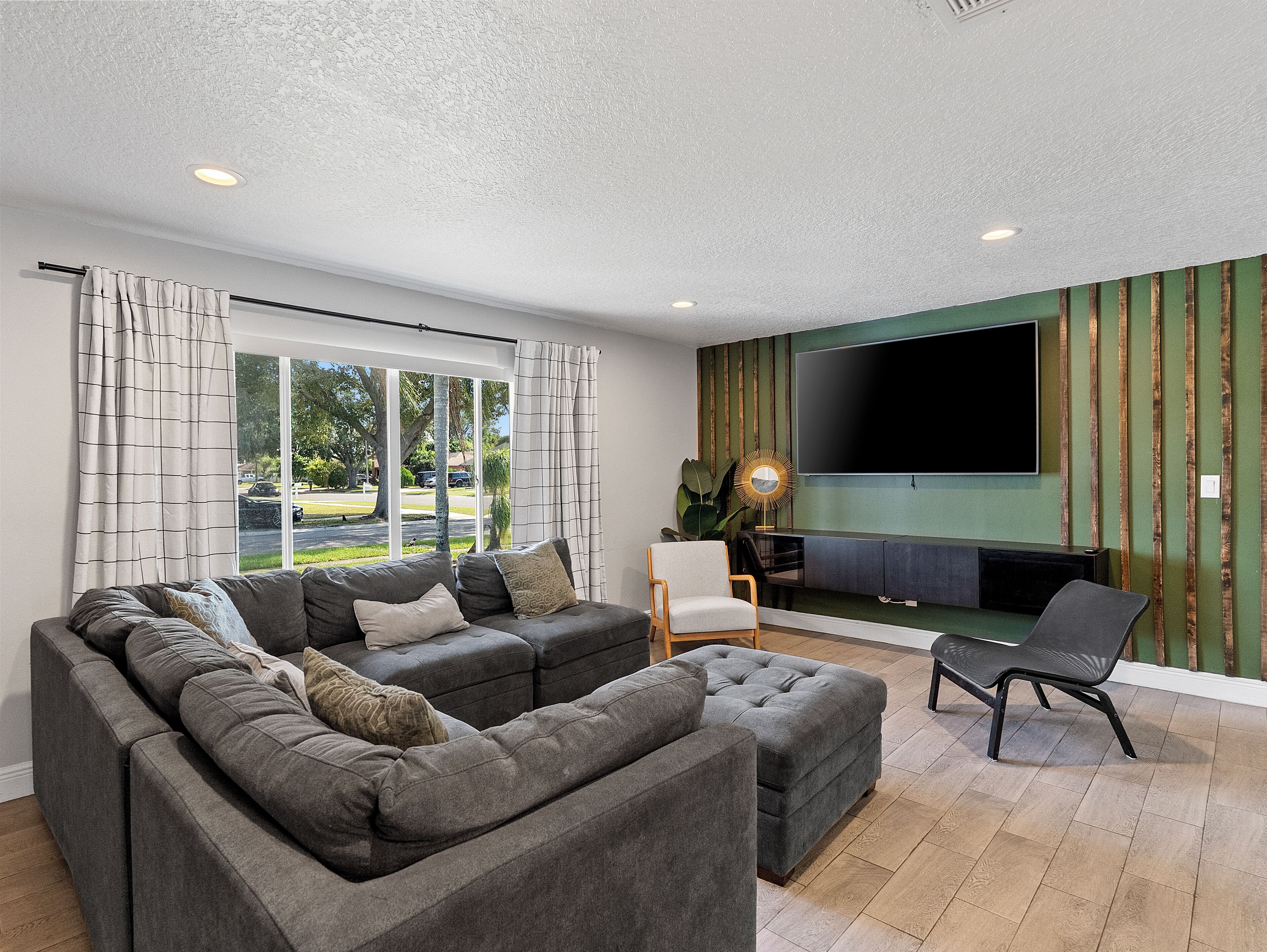 A modern living room with a sectional sofa, a flat-screen TV mounted on a green wall, and a large window with curtains.