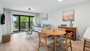 A modern living room with a round wooden dining table, grey cushioned chairs, a grey sofa, and a balcony with a view of greenery. - Beautiful Updates - Relaxing Views - Close to Pool (Naples)