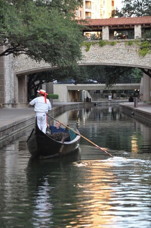 A gondola with a person rowing on a canal, a stone bridge in the background, and a building with a tiled roof.
