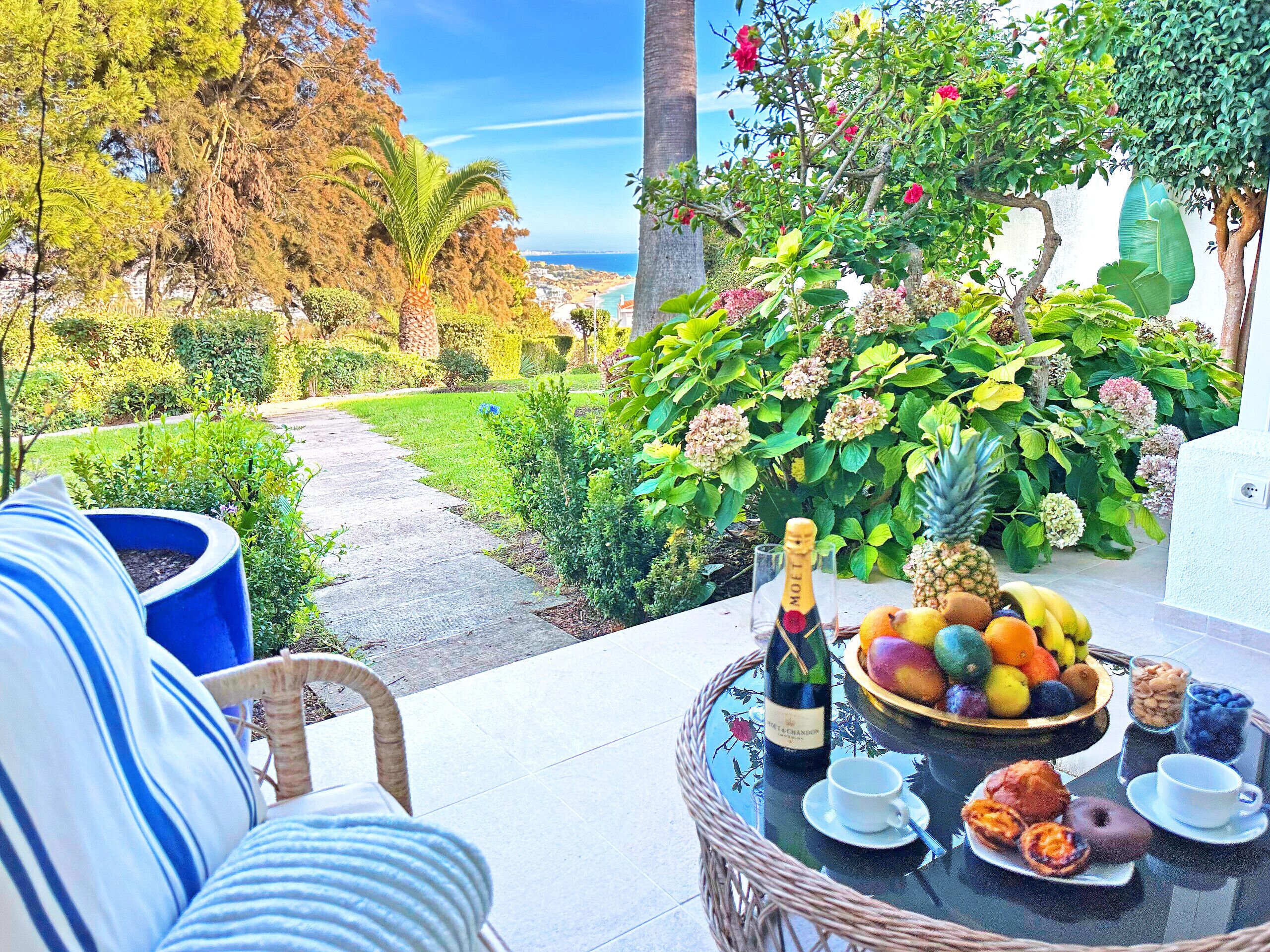 A patio with a wicker chair, a table with a bottle of wine, fruit, pastries, and a view of a garden and ocean.