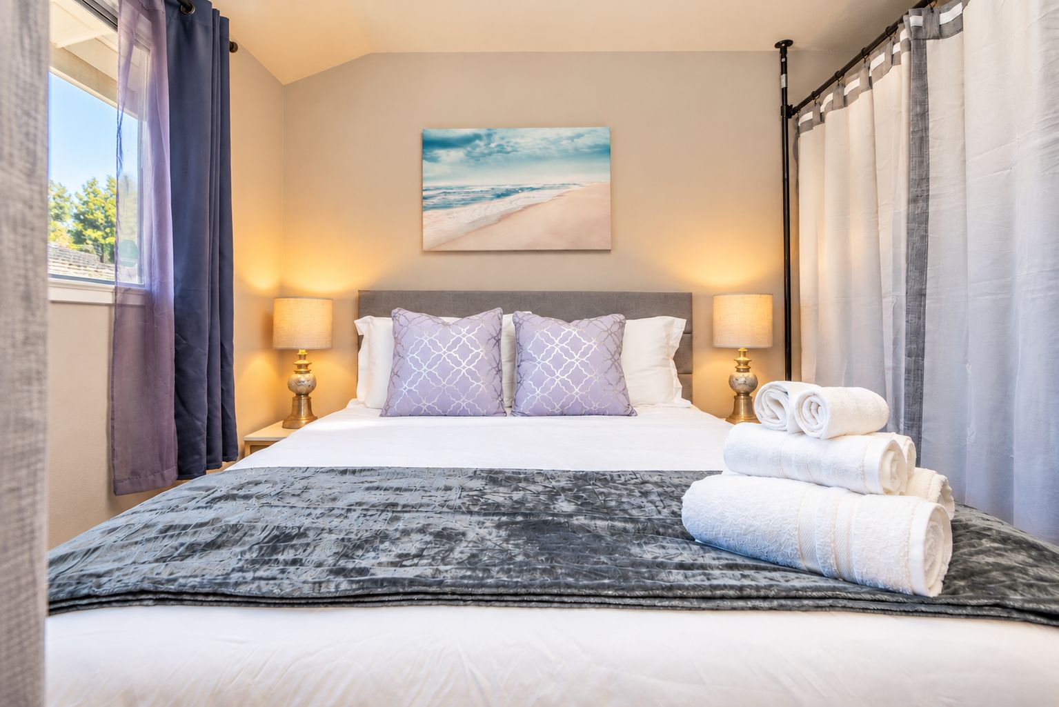 Habitación de hotel con una cama, dos lámparas de noche, un cuadro de una playa y una ventana con cortinas.