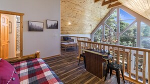 A spacious room with a wooden ceiling, a balcony with a railing, a bed with a plaid blanket, a desk with a computer, and a view of trees through large windows. - Bearfoot Lodge in Angel Fire (Angel Fire)
