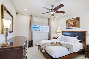 A bedroom with a bed, a ceiling fan, a window with curtains, a bedside table with a lamp, and a painting on the wall. - Beach View Condo in the heart of Pacific Beach! (San Diego)