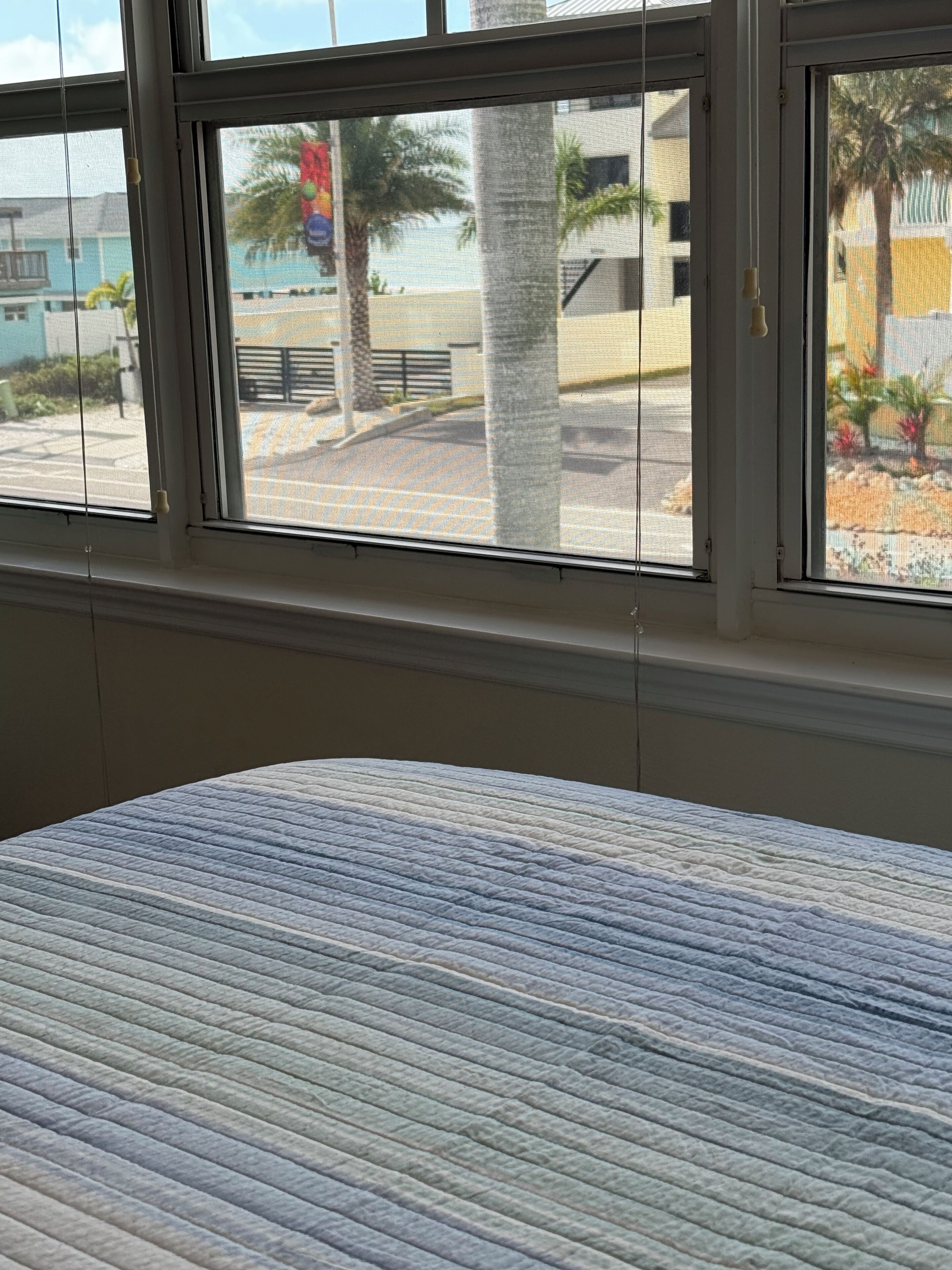 A room with a bed, a window with a view of palm trees and buildings outside.