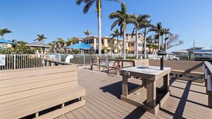 A wooden deck with tables and chairs, palm trees, and a clear blue sky. - Cozy Barefoot Beach Condo E-217 – Just 100 Steps from Indian Shores Beach. (Indian Shores)