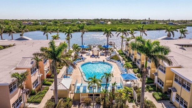A hotel with a central swimming pool surrounded by palm trees and lounge chairs, with a view of a body of water and greenery in the background. - Cozy Barefoot Beach Condo E-217 – Just 100 Steps from Indian Shores Beach. (Indian Shores)