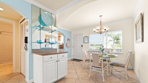 A kitchen with white cabinets, a granite countertop, and a dining area with wicker chairs and a chandelier. - Cozy Barefoot Beach Condo E-217 – Just 100 Steps from Indian Shores Beach. (Indian Shores)